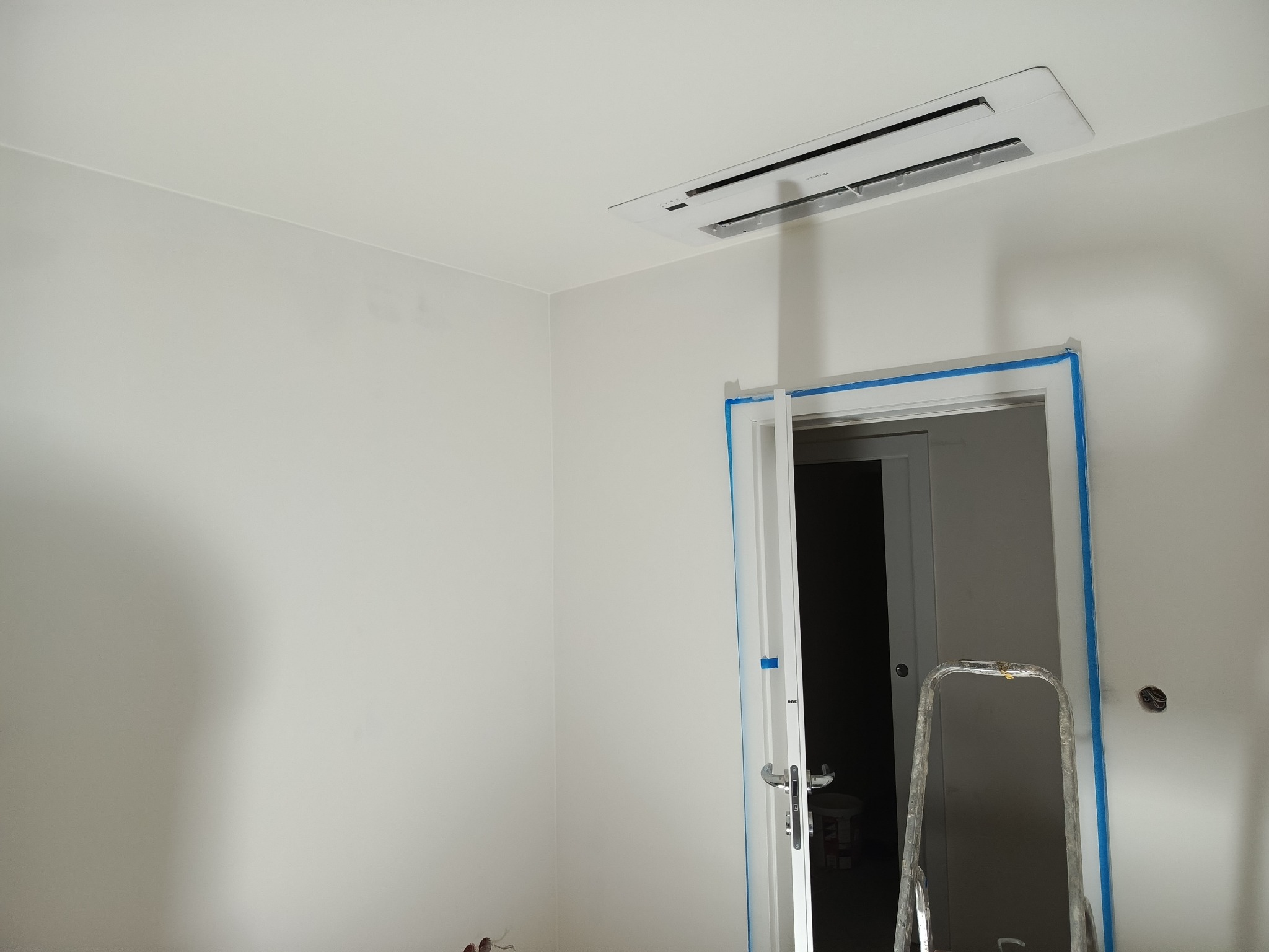 Białe wnętrze po malowaniu z klimatyzatorem kasetonowym i zabezpieczonymi taśmą drzwiami. Widoczna drabina malarska.