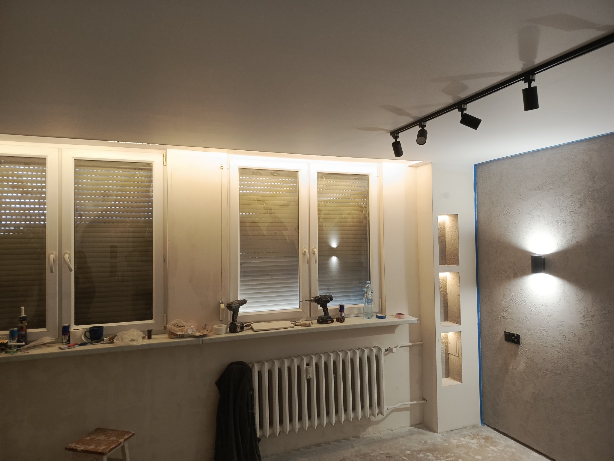 Wnętrze w trakcie remontu: okno z narzędziami, grzejnik, oświetlenie szynowe i dekoracyjna ściana z wnękami. Widoczne ślady prac wykończeniowych.