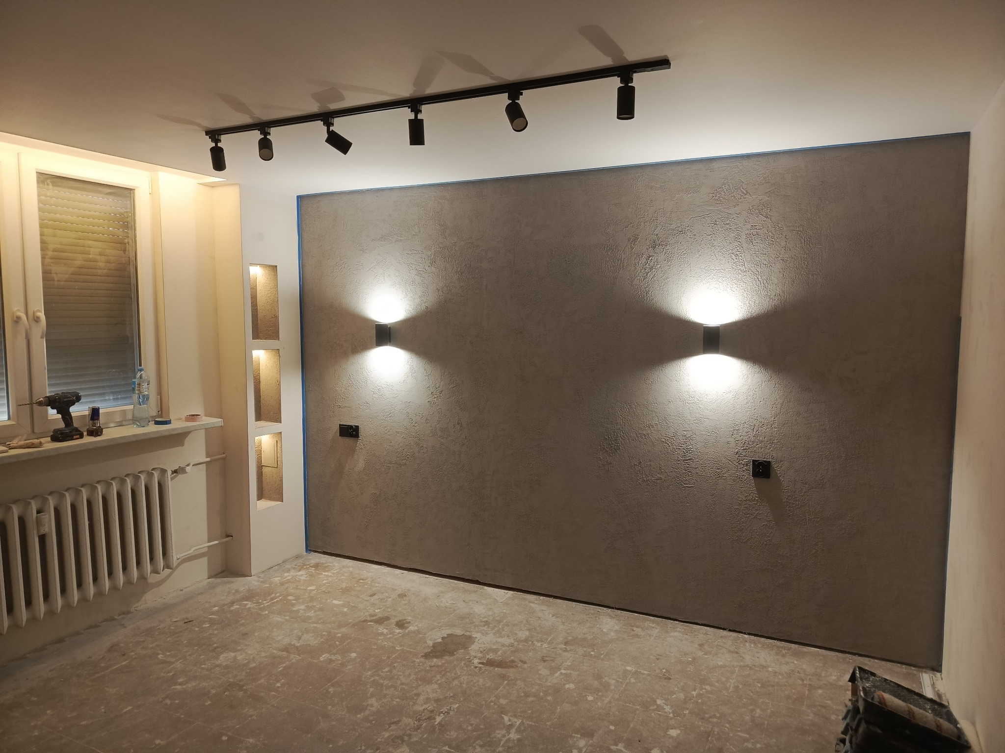 Wykończone wnętrze pokoju z dekoracyjną ścianą w odcieniach szarości, oświetloną przez dwa kinkiety i reflektory sufitowe. Widoczny grzejnik i okno.