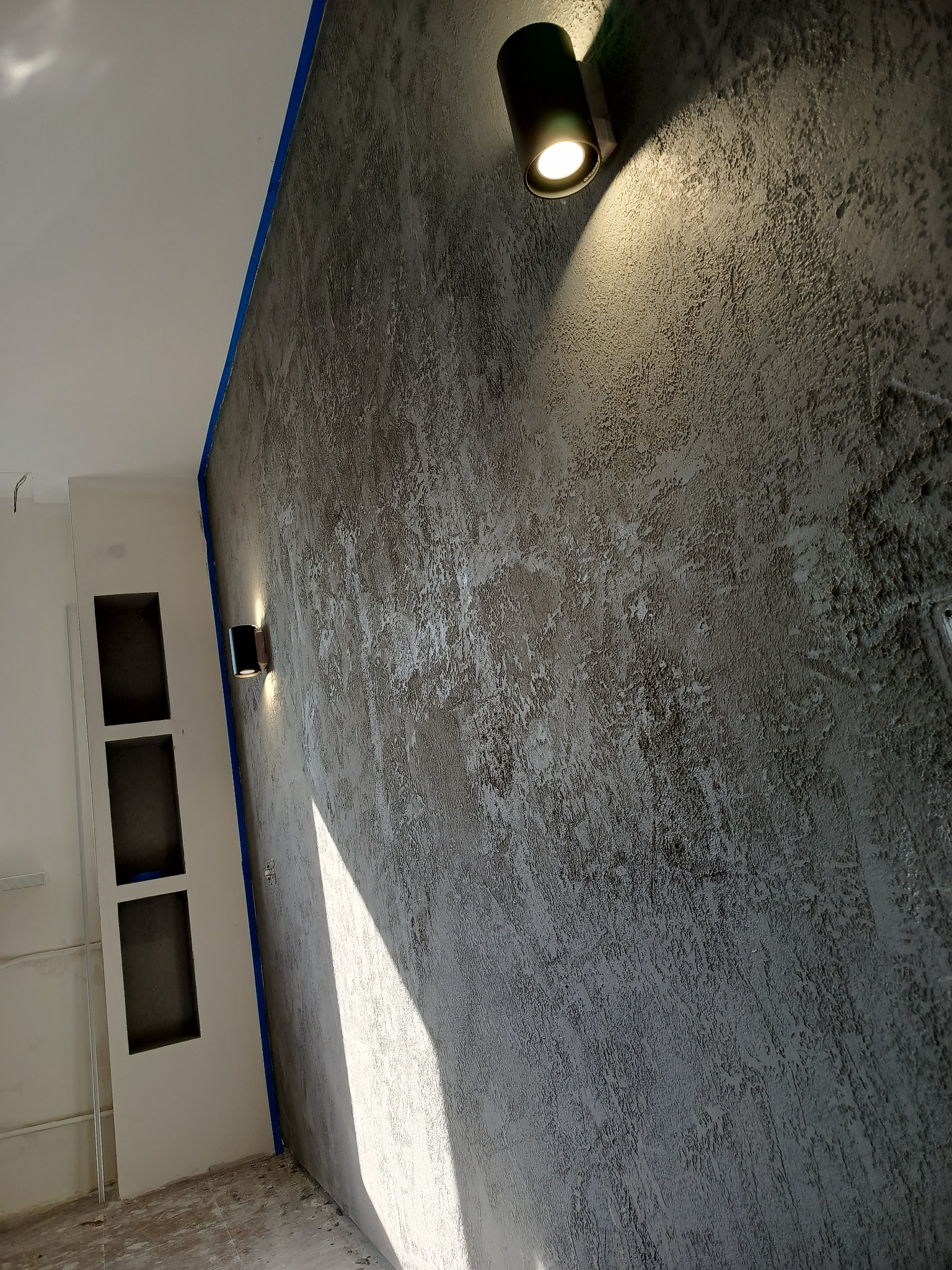 Dekoracyjna ściana z efektem betonu architektonicznego, oświetlona dwoma nowoczesnymi lampami ściennymi. Widoczny fragment ściany z wnękami.