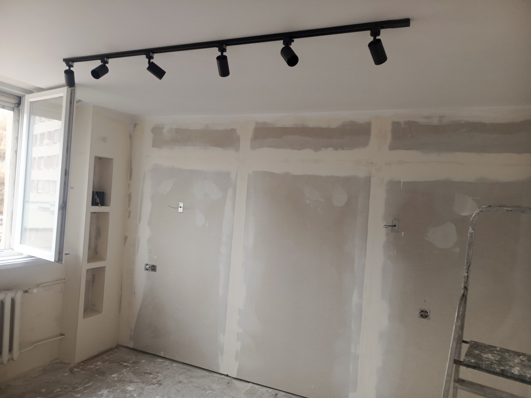 Pokój w trakcie szpachlowania ścian z płyt karton-gips, widoczne puszki elektryczne, drabina i oświetlenie szynowe. Okno z grzejnikiem.