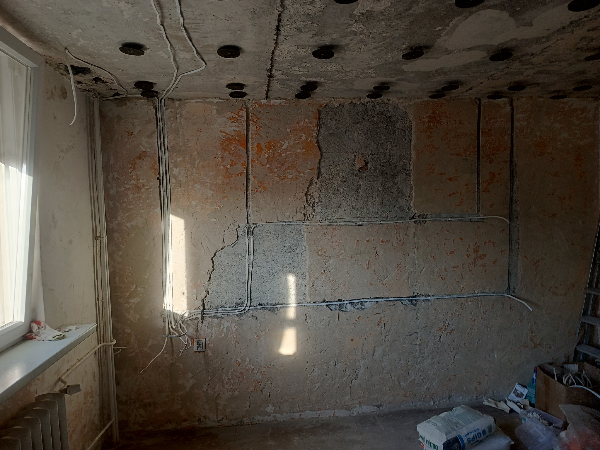 Pokój w trakcie remontu: ściany z resztkami starej farby, widoczne przewody elektryczne, sufit z zamontowanymi puszkami. Worki z gipsem na podłodze.