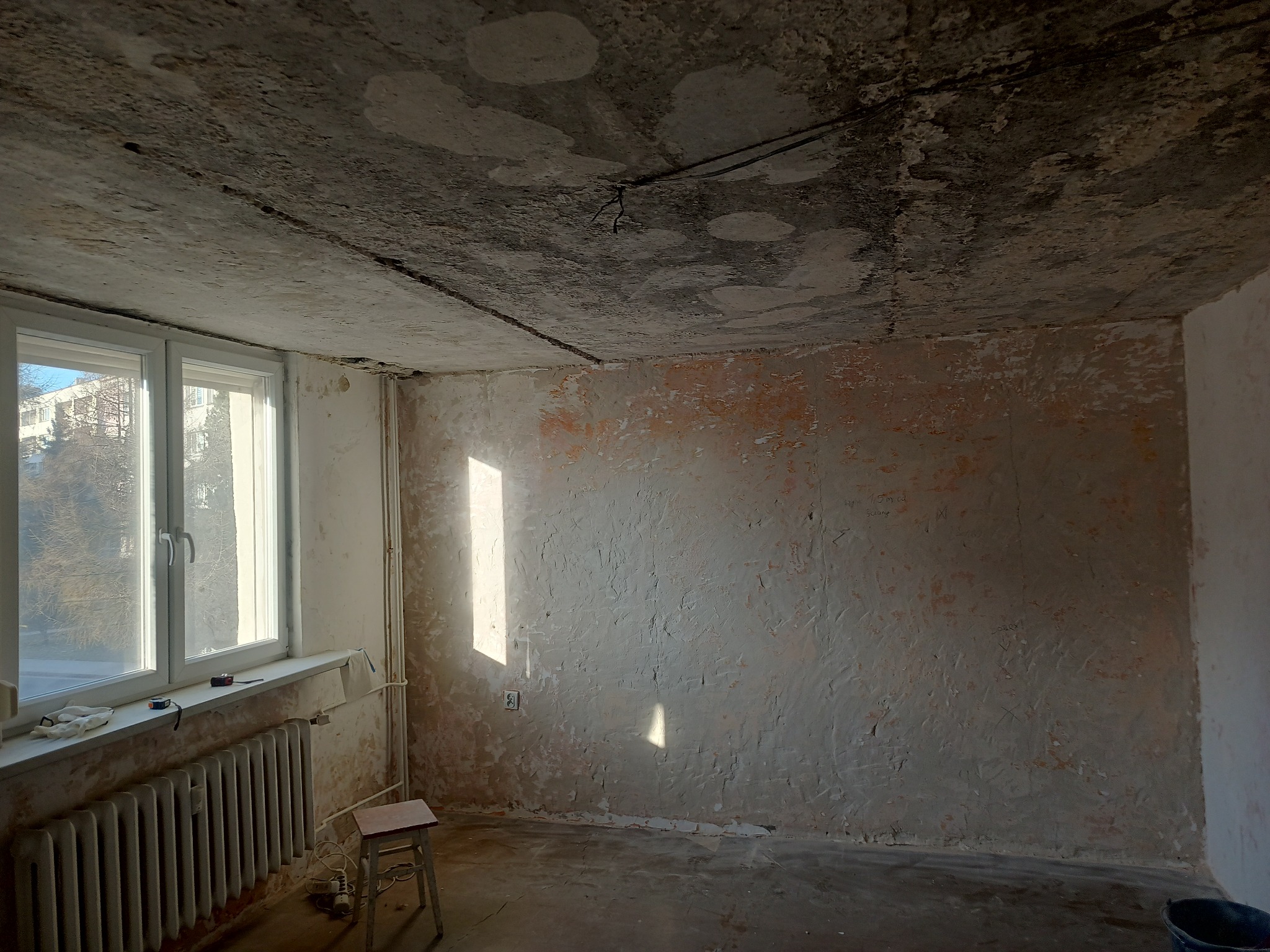 Pokój w trakcie remontu w Bytomiu: surowy strop, ściany z resztkami farby, okno z grzejnikiem, mały stołek i wiadro. Przygotowanie do malowania lub szpachlowania.