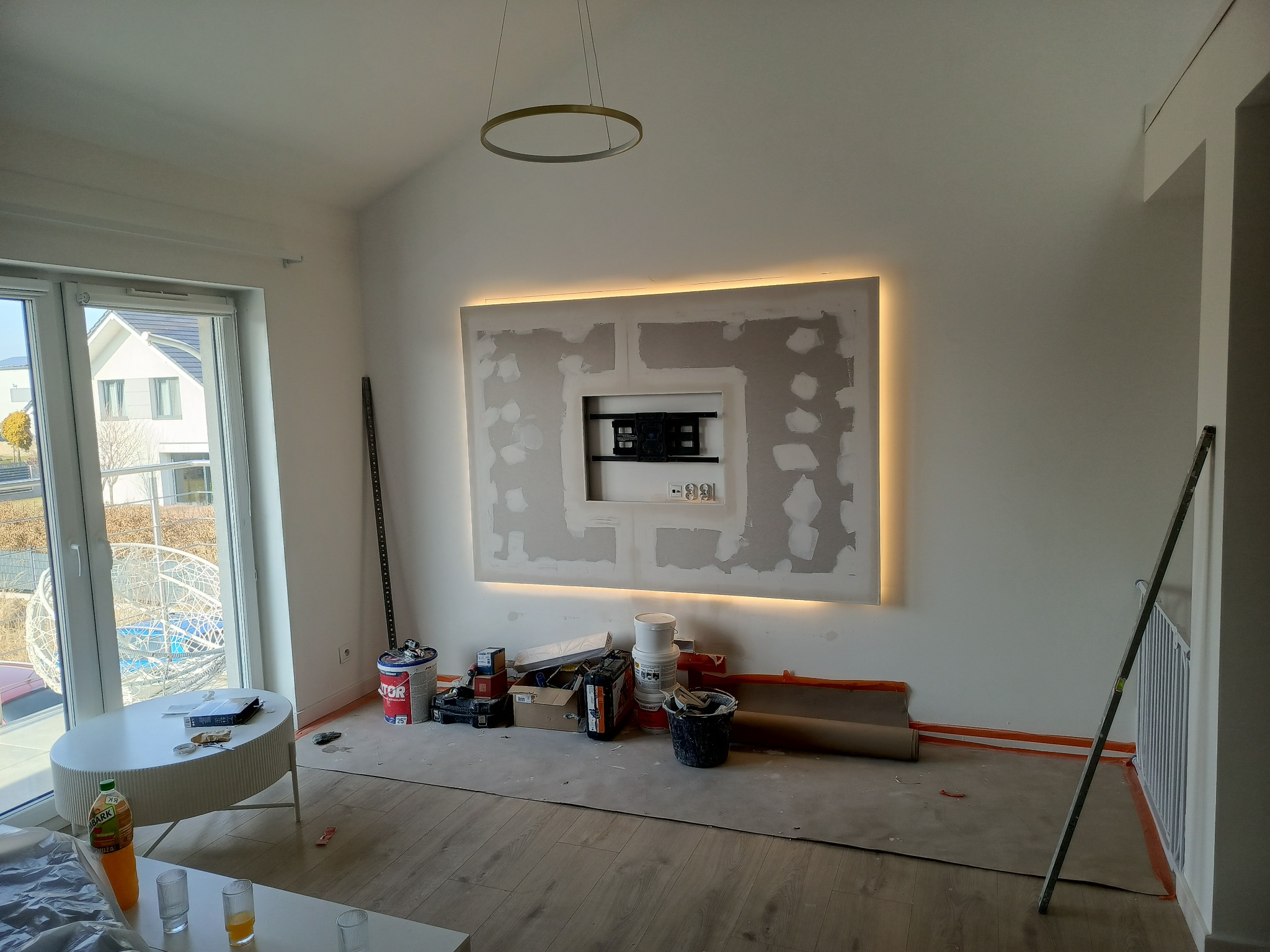 Ściana z płyt gipsowych w trakcie szpachlowania, podświetlona taśmą LED. Widoczne narzędzia i materiały budowlane. W tle okno z widokiem na zewnątrz.