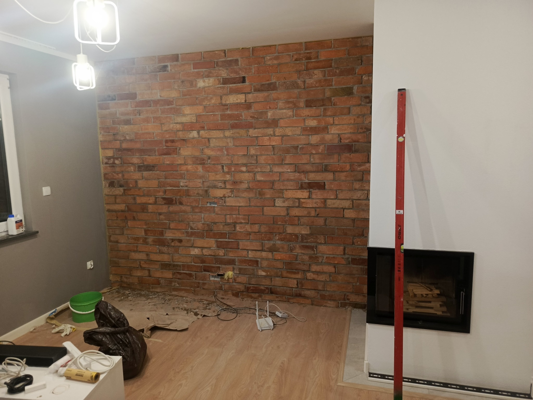Ściana z cegły w trakcie wykończenia wnętrza, widoczny poziomica przy kominku, elementy instalacji elektrycznej i narzędzia. Oświetlenie sufitowe.