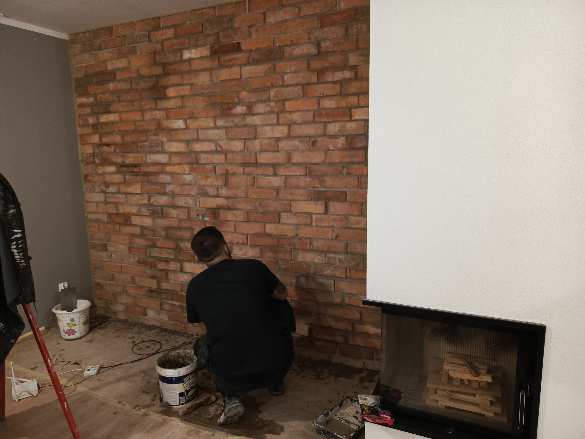 Pracownik w trakcie układania cegieł na ścianie, widoczne narzędzia i materiały budowlane. Fragment ściany pomalowany na biało, obok kominek.