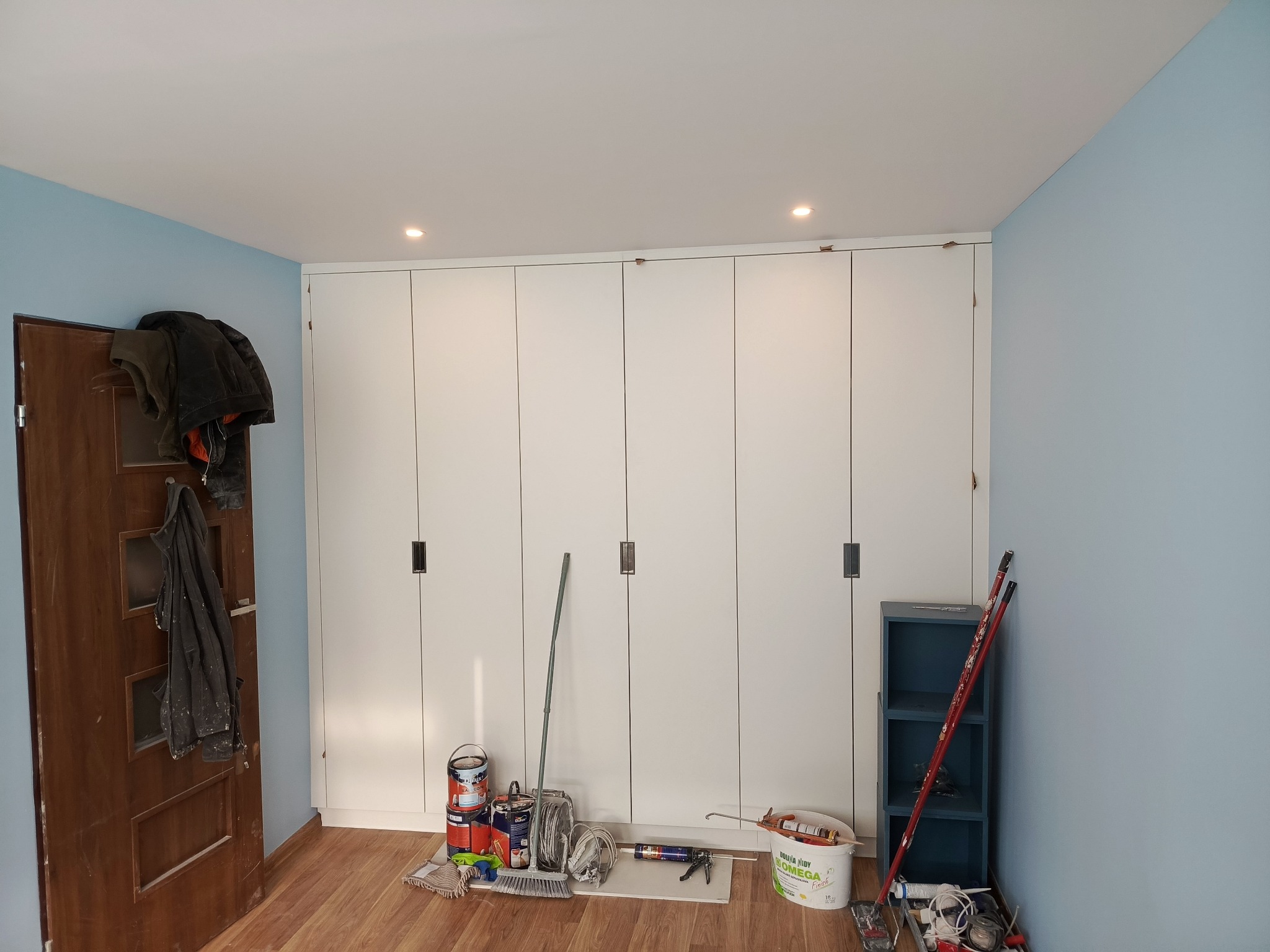 Błękitne ściany i biała zabudowa szafy wnękowej. Na podłodze widoczne narzędzia i materiały malarskie, sugerujące etap wykończeniowy prac remontowych.