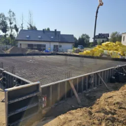 Zbrojenie fundamentu pod budynek jednorodzinny, widoczne szalunki i siatka zbrojeniowa. W tle dom w trakcie budowy i pompa do betonu.