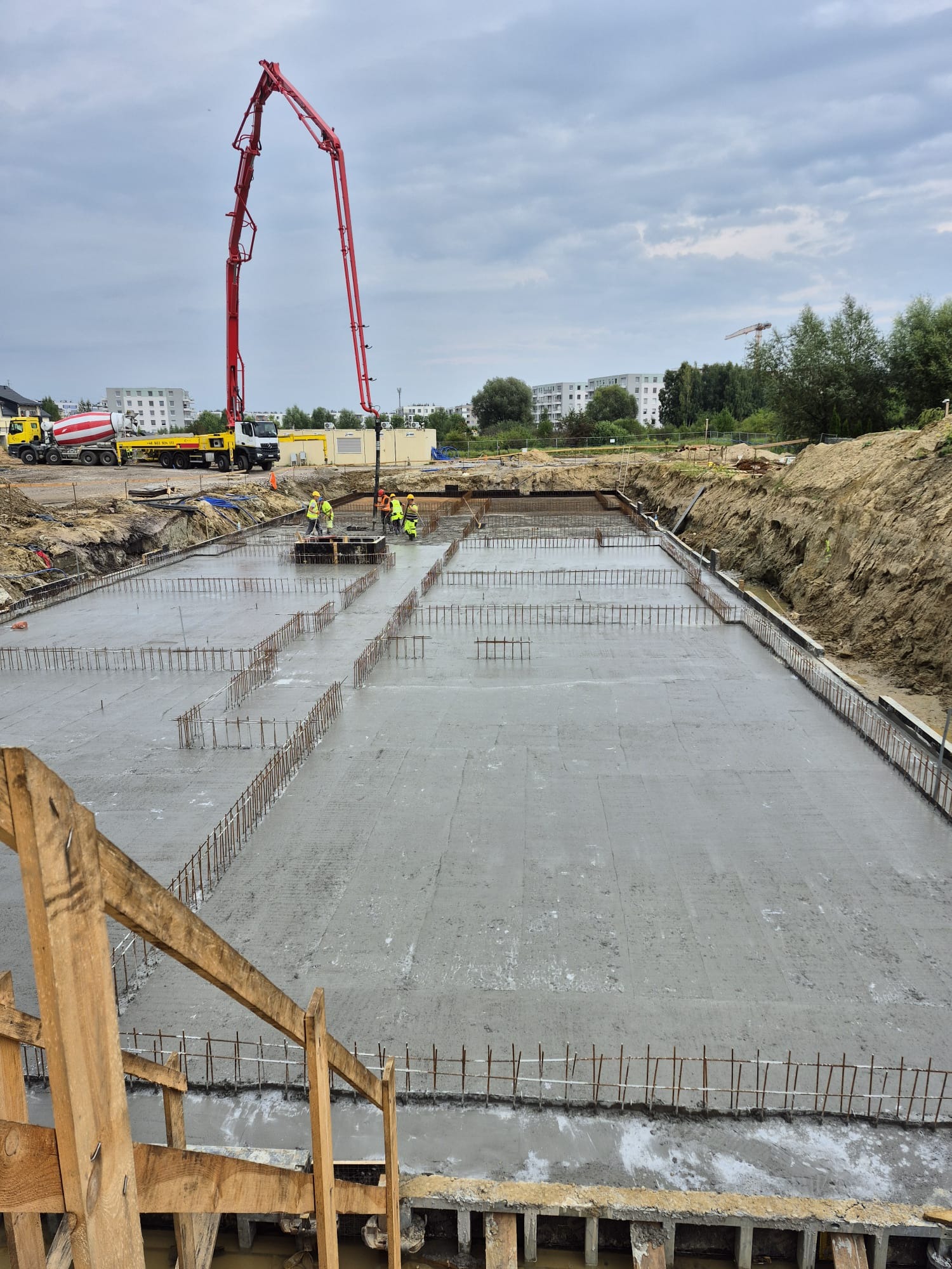 Wylewka fundamentu zbrojonego, widoczna pompa do betonu i betoniarka. Prace budowlane w toku, teren budowy, budynki w tle.