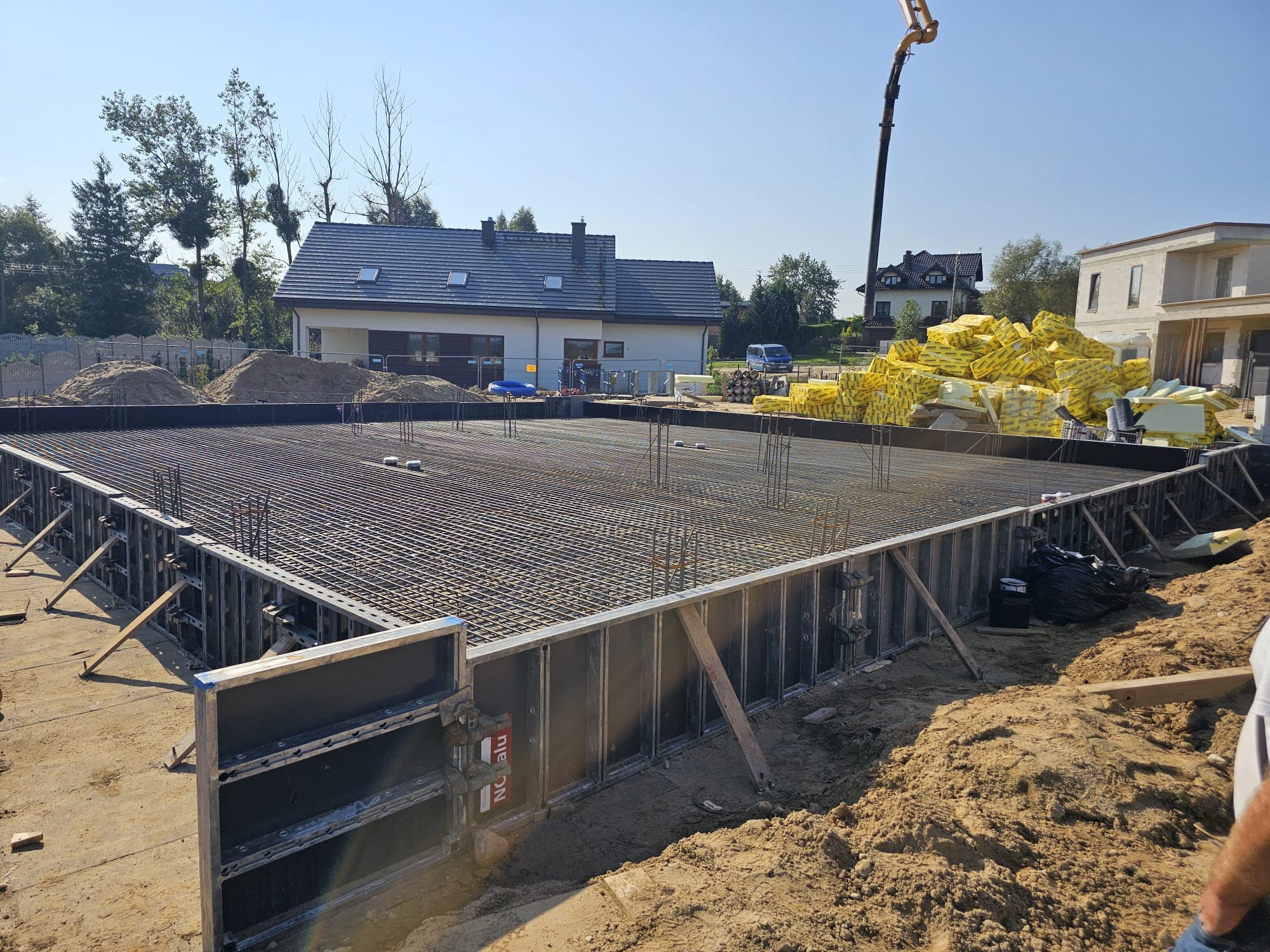 Zbrojenie fundamentu pod budynek jednorodzinny, widoczne szalunki i siatka zbrojeniowa. W tle dom w trakcie budowy i pompa do betonu.