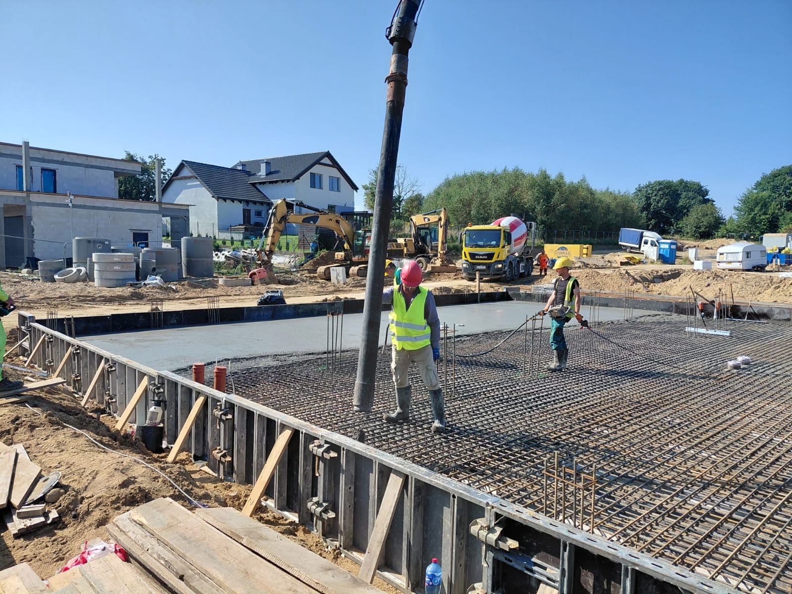 Wylewanie betonu na przygotowane zbrojenie fundamentów pod budowę domu jednorodzinnego. Na budowie pracują robotnicy, w tle betoniarka i koparki.