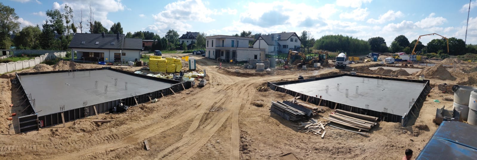 Panorama z budowy domu: dwa wylane fundamenty z wystającymi prętami, teren budowy z maszynami i materiałami, w tle domy jednorodzinne i zielone drzewa.