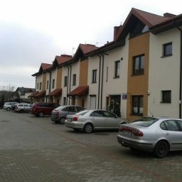 Marcin - Rząd szeregowych domów z czerwonymi dachami i beżowo-brązowymi elewacjami, zaparkowane samochody na brukowanym podjeździe, pochmurne niebo w tle.