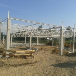 Marcin - Konstrukcja stalowa hali w trakcie montażu, widoczne betonowe słupy i elementy kratownicowe. Teren budowy z piaskiem i sprzętem budowlanym w tle.
