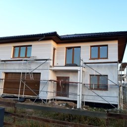 Marcin - Dwukondygnacyjny dom w trakcie prac wykończeniowych elewacji, z widocznymi rusztowaniami i brązowymi oknami. Częściowo otynkowany, z ciemnym dachem i drewnianym ogrodzeniem.