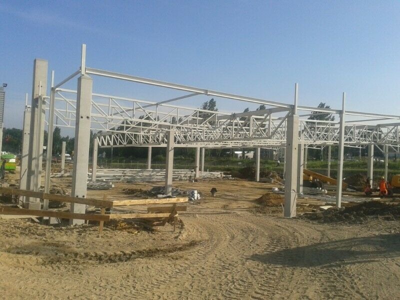 Konstrukcja stalowa hali w trakcie montażu, widoczne betonowe słupy i elementy kratownicowe. Teren budowy z piaskiem i sprzętem budowlanym w tle.