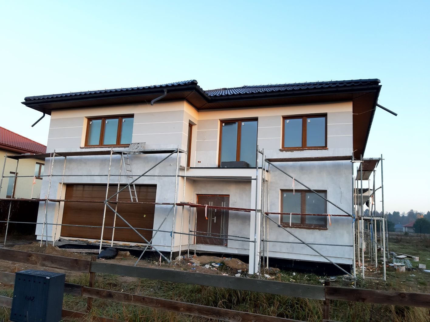Dwukondygnacyjny dom w trakcie prac wykończeniowych elewacji, z widocznymi rusztowaniami i brązowymi oknami. Częściowo otynkowany, z ciemnym dachem i drewnianym ogrodzeniem.
