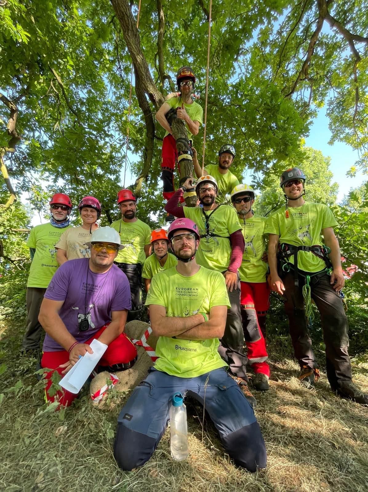Grupa arborystów w kaskach i strojach roboczych pozuje na tle drzewa. Jeden z nich wspina się po pniu. Widoczne liny i sprzęt do arborystyki.