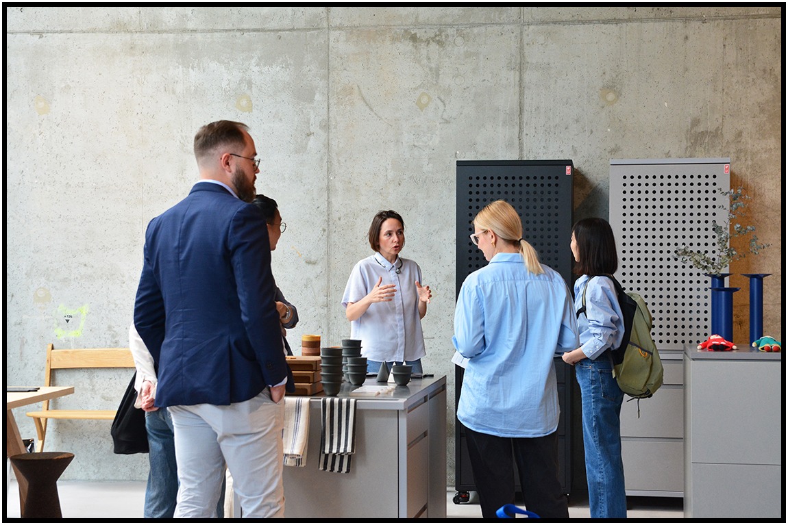 Prezentacja naczyń ceramicznych w loftowym wnętrzu. Osoby słuchają kobiety gestykulującej przy stole z naczyniami. W tle betonowa ściana i metalowe szafki.