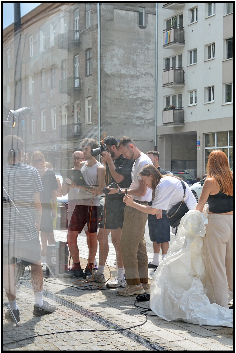 Ekipa fotograficzna w plenerze podczas sesji zdjęciowej. W tle budynki, widoczne oświetlenie studyjne, fotografowie z aparatami i asystenci. Ujęcie przez szybę.