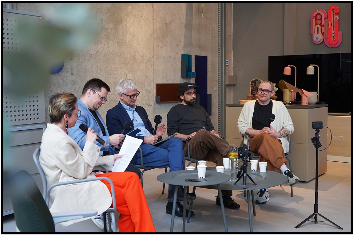 Panel dyskusyjny z mikrofonami, nagrywany kamerą na statywie. W tle betonowa ściana i designerskie lampki.