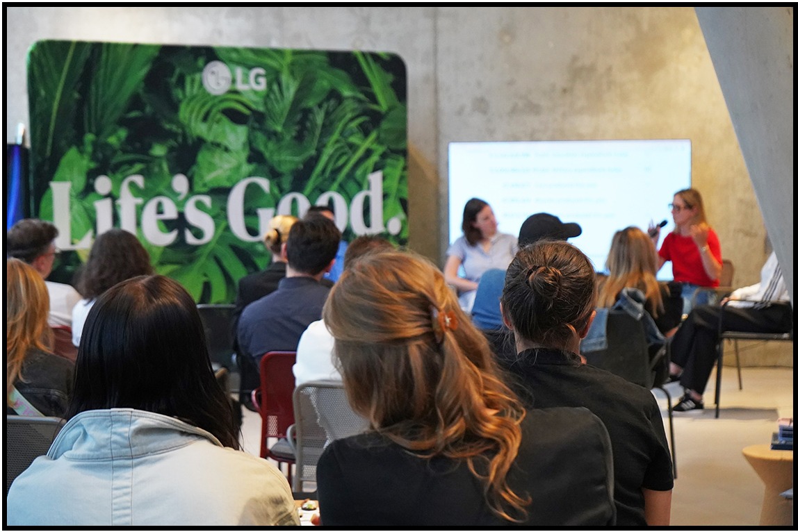 Widok zza pleców na prelegentki i publiczność podczas prelekcji z logo LG w tle. Kobieta w czerwonej bluzce gestykuluje, a druga siedzi obok na krześle.