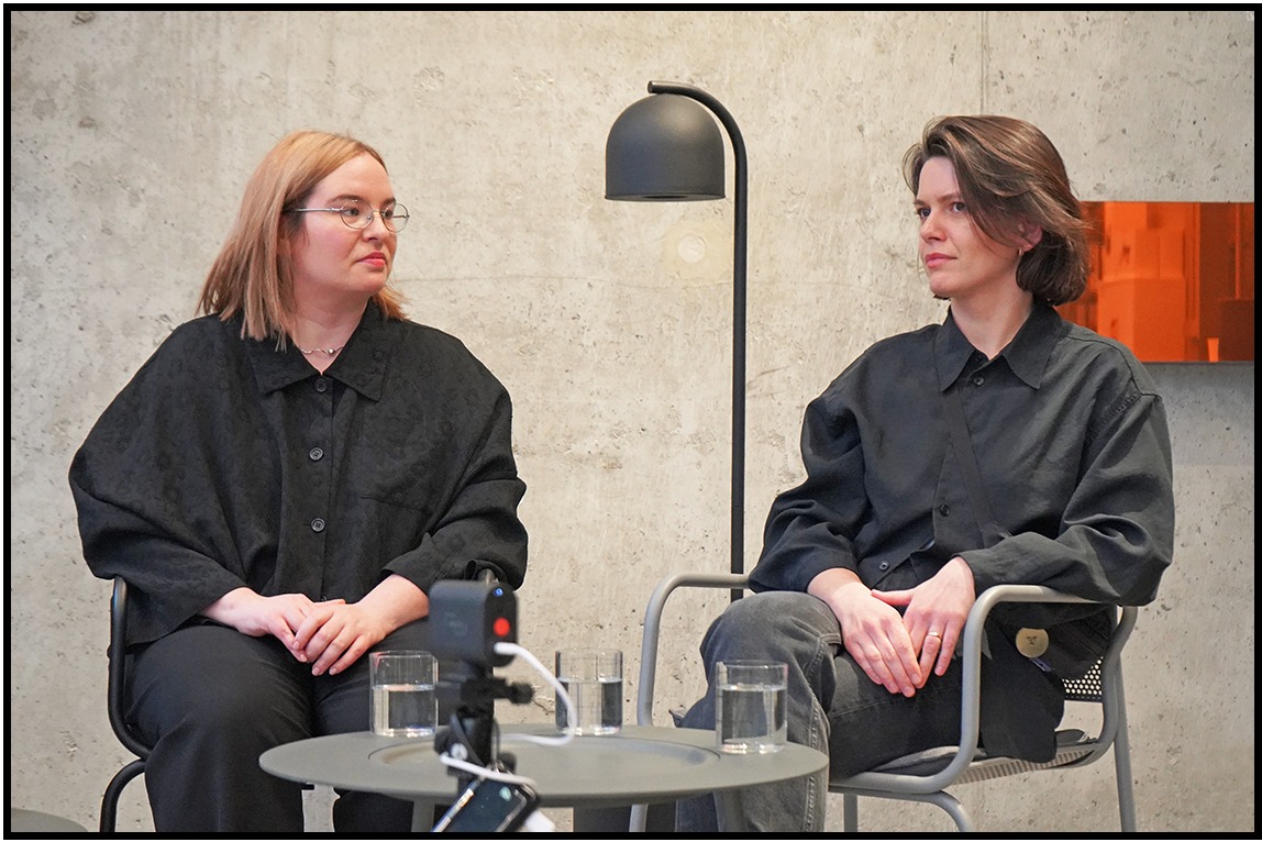 Dwie kobiety w ciemnych koszulach siedzą na krzesłach przy stoliku z wodą. W tle betonowa ściana i lampa. Ujęcie z kamery podczas wywiadu.