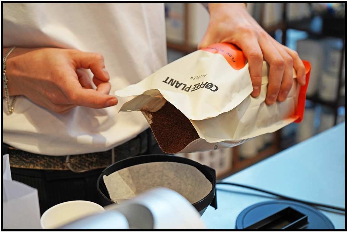 Nalewanie kawy z paczki 'Coffee Plant' do filtra. Widoczne ręce, paczka z kawą, filtr, biała koszula i srebrna bransoletka na nadgarstku. Ujęcie z bliska.