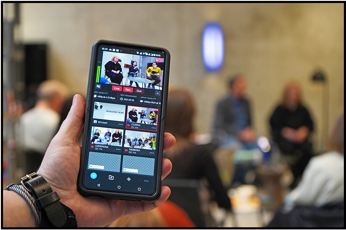 Smartfon w dłoni wyświetla podgląd transmisji na żywo z panelu dyskusyjnego. W tle rozmyte sylwetki publiczności. Widoczny zegarek na ręce.