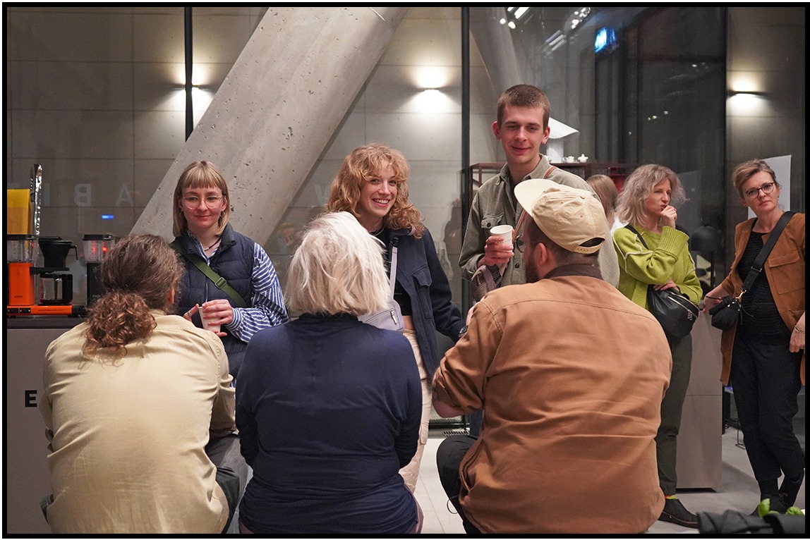 Grupa osób w jasnym wnętrzu, część siedzi, część stoi, rozmawiają i piją kawę. Widoczny ekspres do kawy, betonowy filar i duże okno.