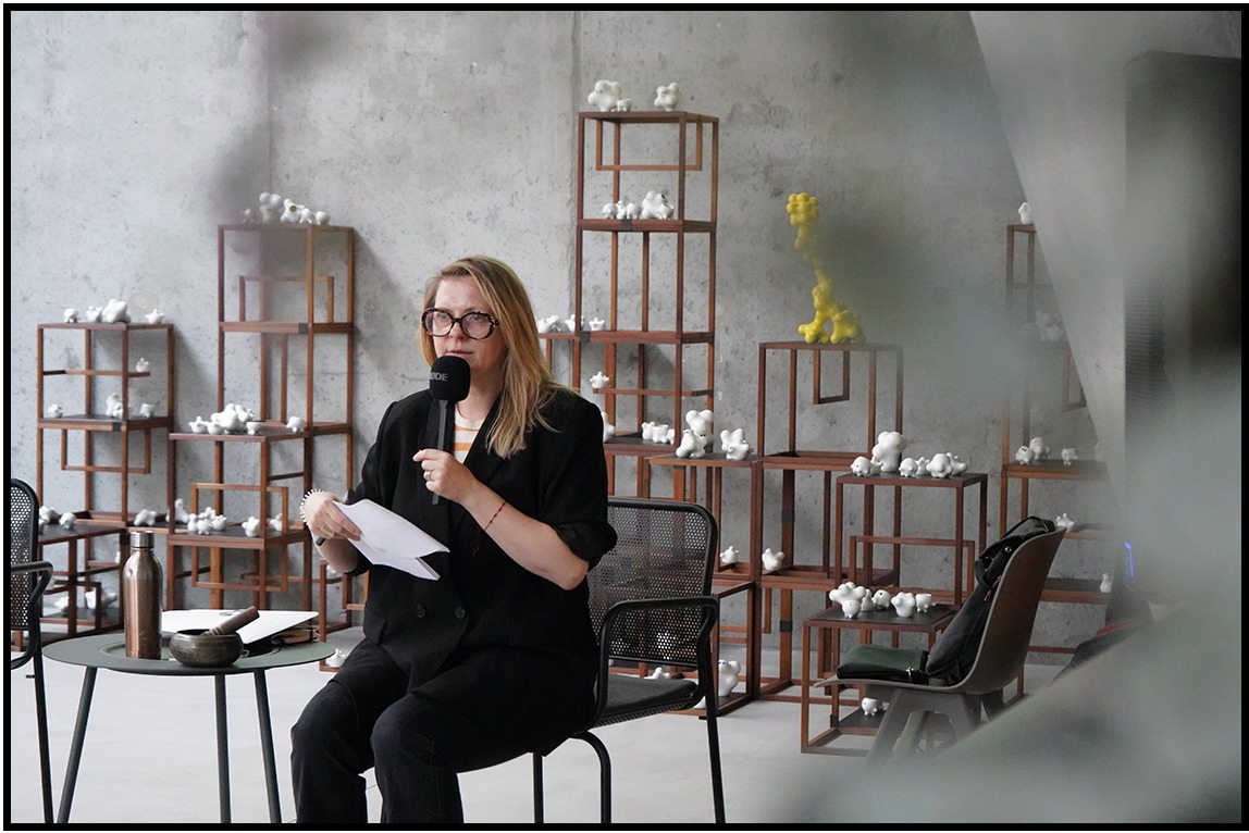 Kobieta z mikrofonem przemawia na tle betonowej ściany i drewnianych regałów z białymi figurkami. W tle widoczna żółta rzeźba. Ubrana w czarny żakiet i okulary.