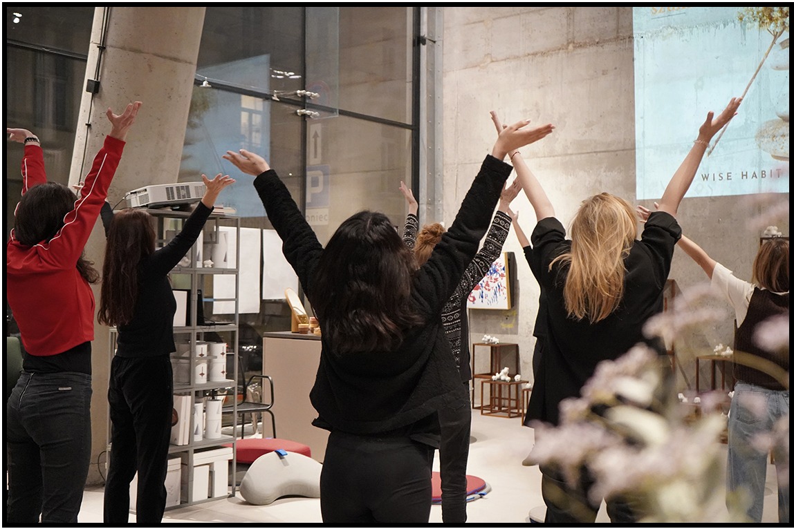 Grupa kobiet z podniesionymi rękoma stoi tyłem do widza w jasnym pomieszczeniu z projektorem wyświetlającym napis 'WISE HABIT'.