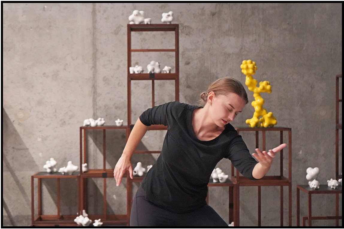 Kobieta w czarnym stroju pozuje na tle regałów z białymi i żółtymi rzeźbami, tworząc artystyczną kompozycję w szarej przestrzeni.