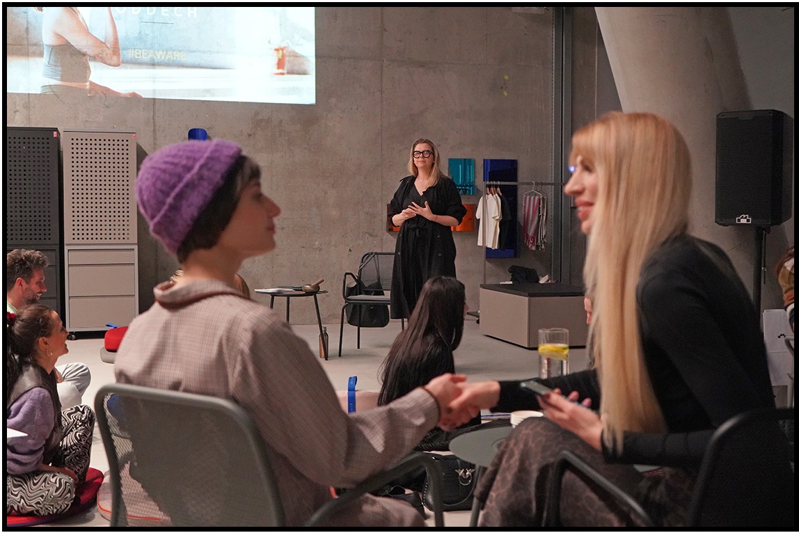 Wnętrze sali konferencyjnej z widocznymi uczestnikami spotkania, prelegentką i rzutnikiem wyświetlającym prezentację. Osoby siedzą na krzesłach i poduszkach.