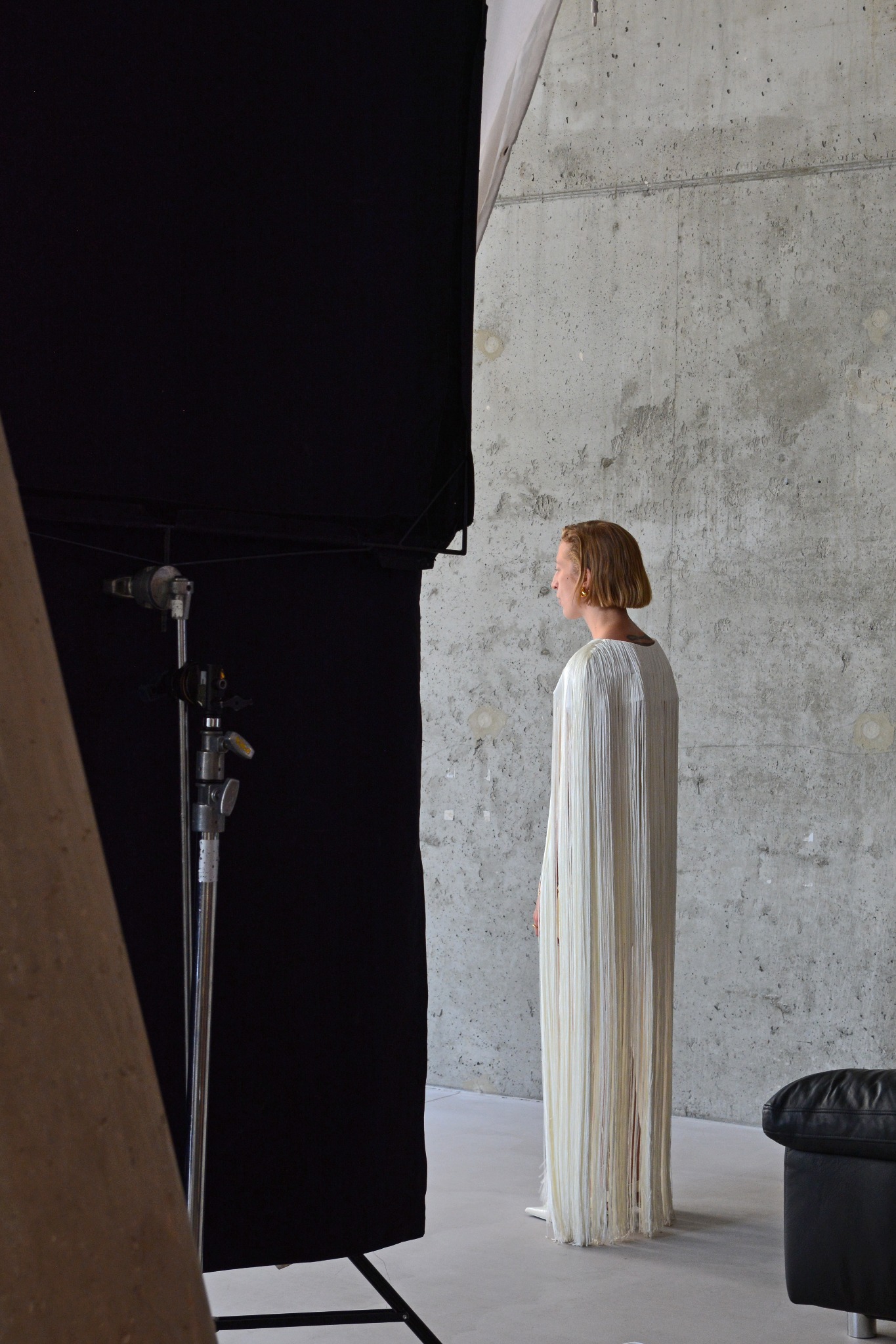 Sesja zdjęciowa w studio w Warszawie: modelka w białej sukni z frędzlami, ustawiona tyłem na tle betonowej ściany, widoczne oświetlenie studyjne i czarny ekran.