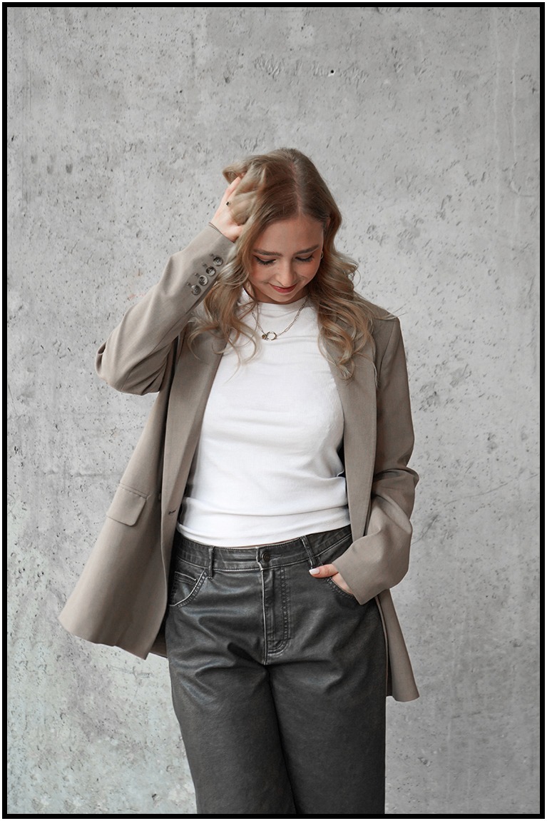 Portret kobiety w beżowym żakiecie i białym topie na tle szarej ściany. Uśmiechnięta, z ręką we włosach, w swobodnej pozie.