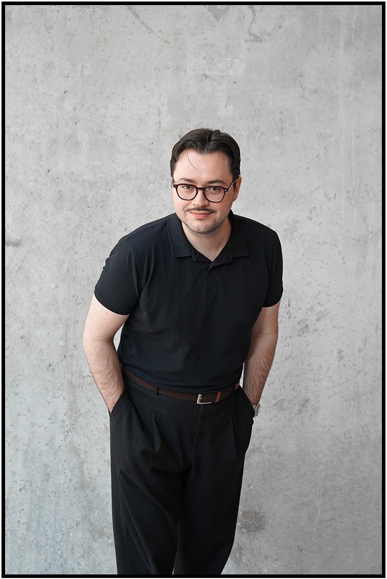 Mężczyzna w okularach i czarnej koszulce polo pozuje na neutralnym tle. Widoczne dłonie w kieszeniach spodni, brązowy pasek i zegarek na ręce.