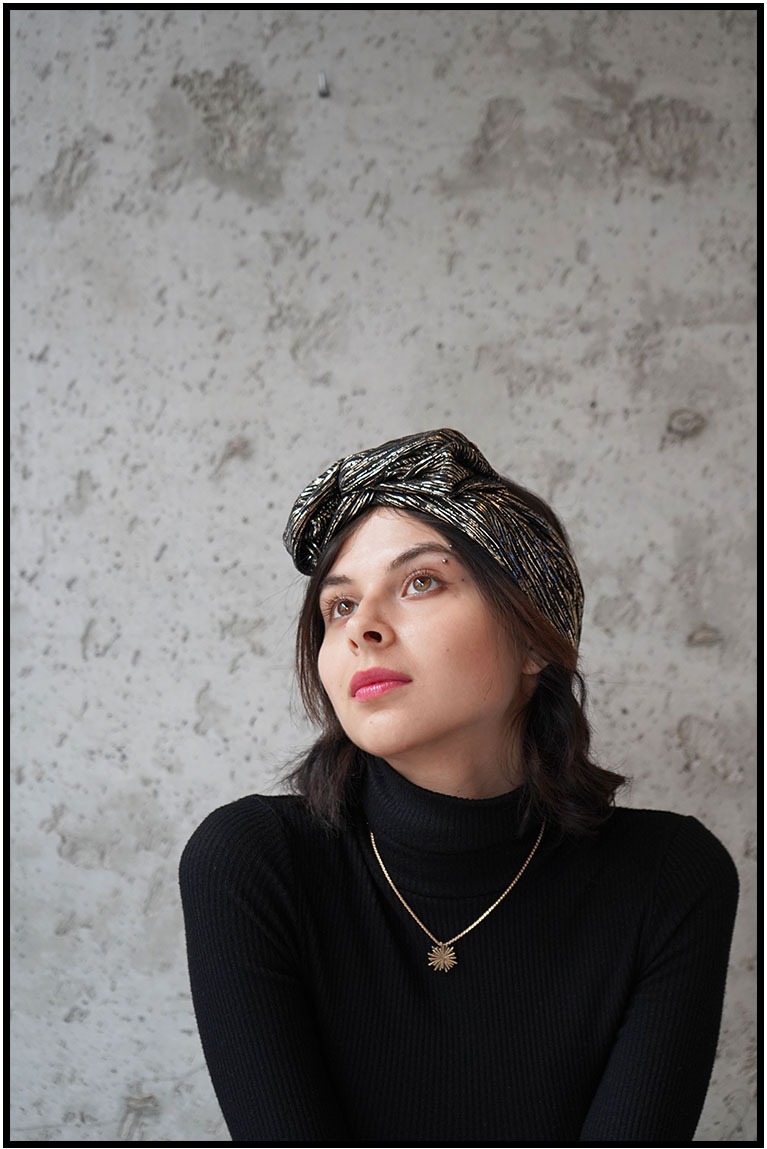 Portret kobiety w czarnym golfie i ozdobnym turbanie, z delikatnym makijażem i złotym naszyjnikiem na tle szarej ściany. Spojrzenie skierowane w górę.