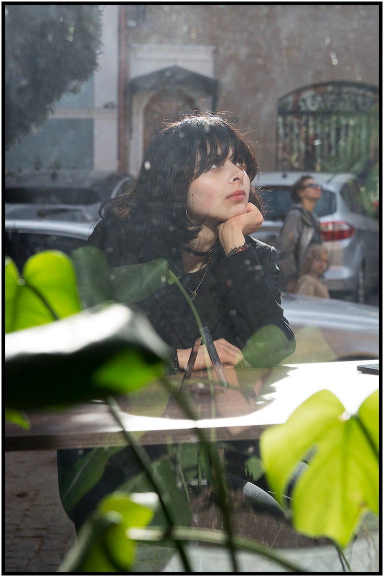 Portret kobiety z ciemnymi włosami, opierającej brodę na dłoni przy stoliku w kawiarni, z widokiem na ulicę przez okno i roślinami w pierwszym planie.