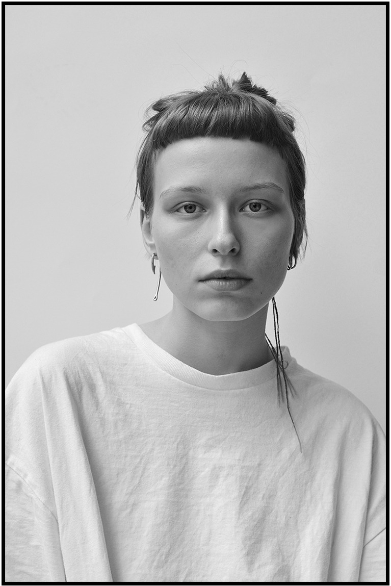 Monochromatyczny portret kobiety z grzywką, w białym T-shircie, z oryginalną biżuterią w uszach, na jasnym tle. Ujęcie z bliska, skupione na twarzy i wyrazie.