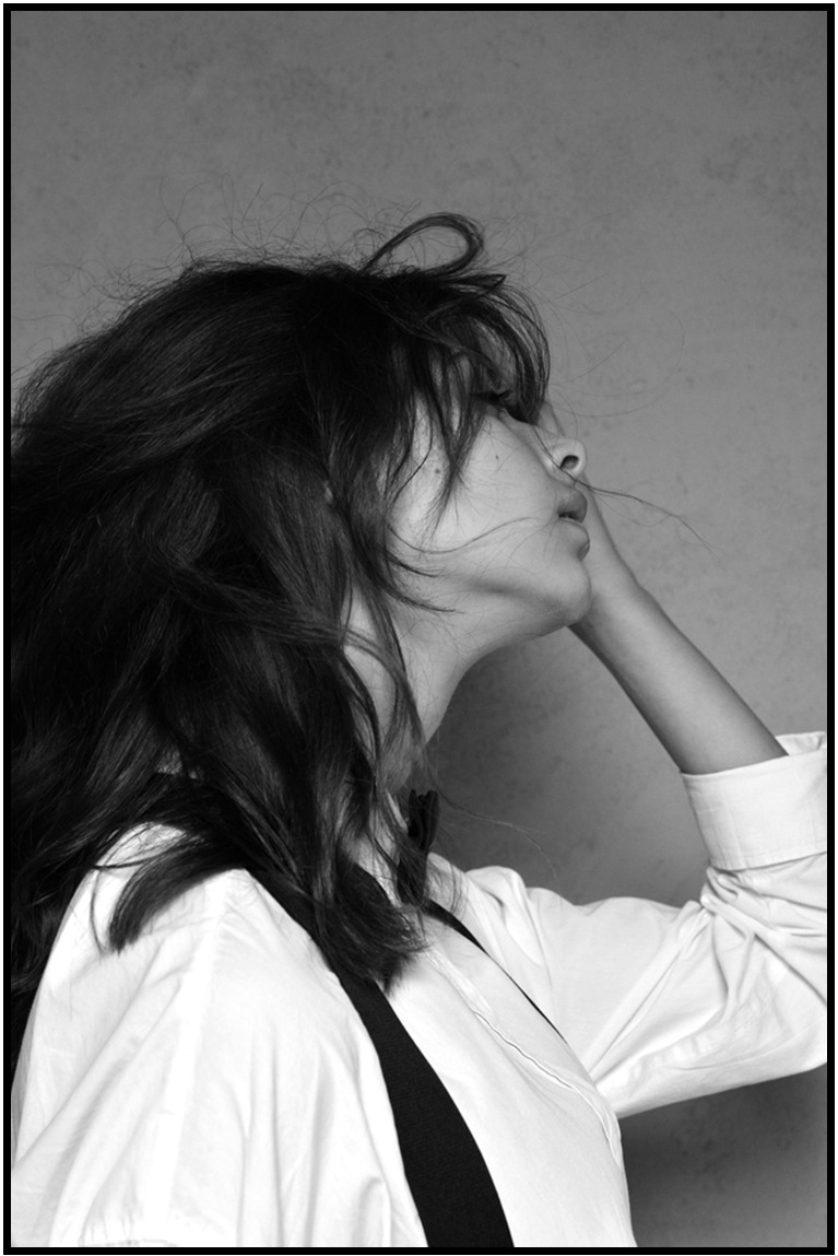 Czarno-biały portret kobiety z rozwianymi włosami, w białej koszuli i szelkach, spoglądającej w górę. Profil, delikatne światło, stonowane tło, artystyczny charakter.
