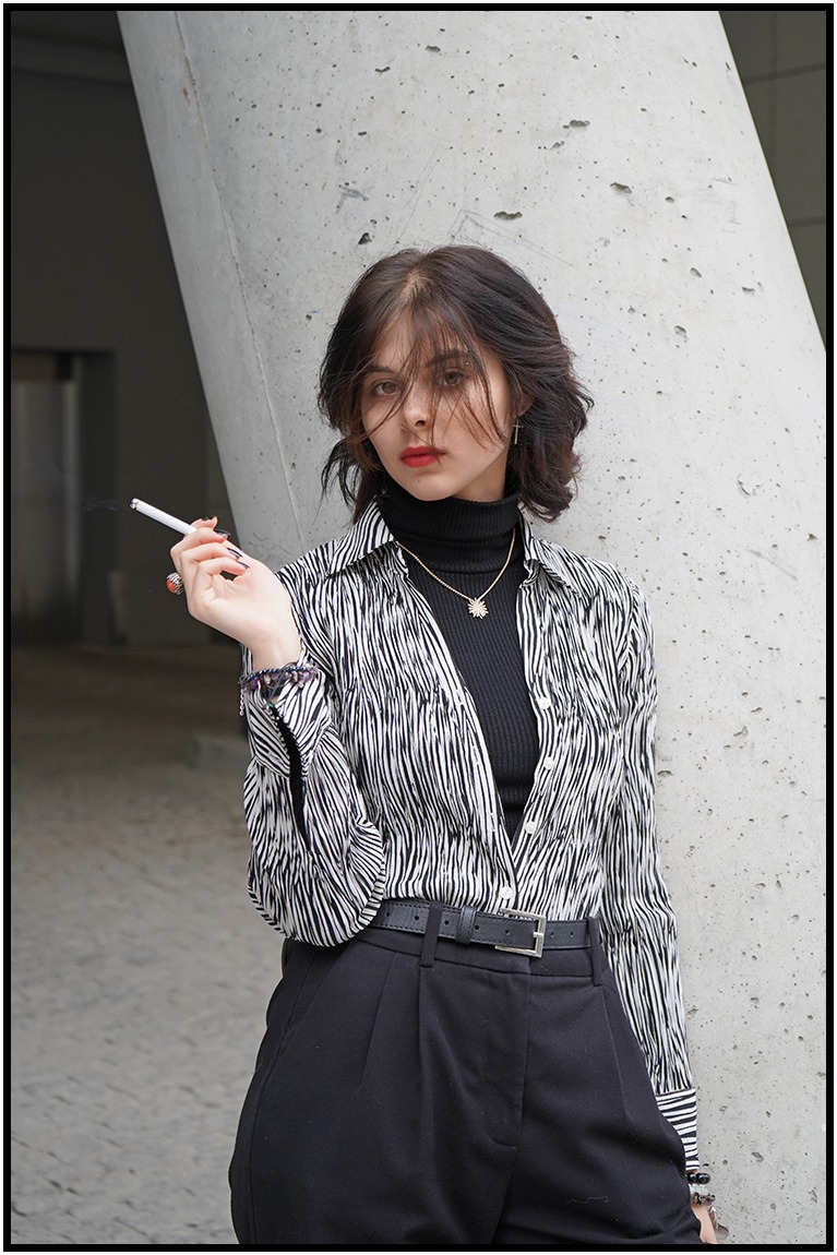 Portret kobiety w czarno-białej koszuli w paski, czarnym golfie i spodniach, opierającej się o betonowy filar z papierosem w dłoni, na zewnątrz.