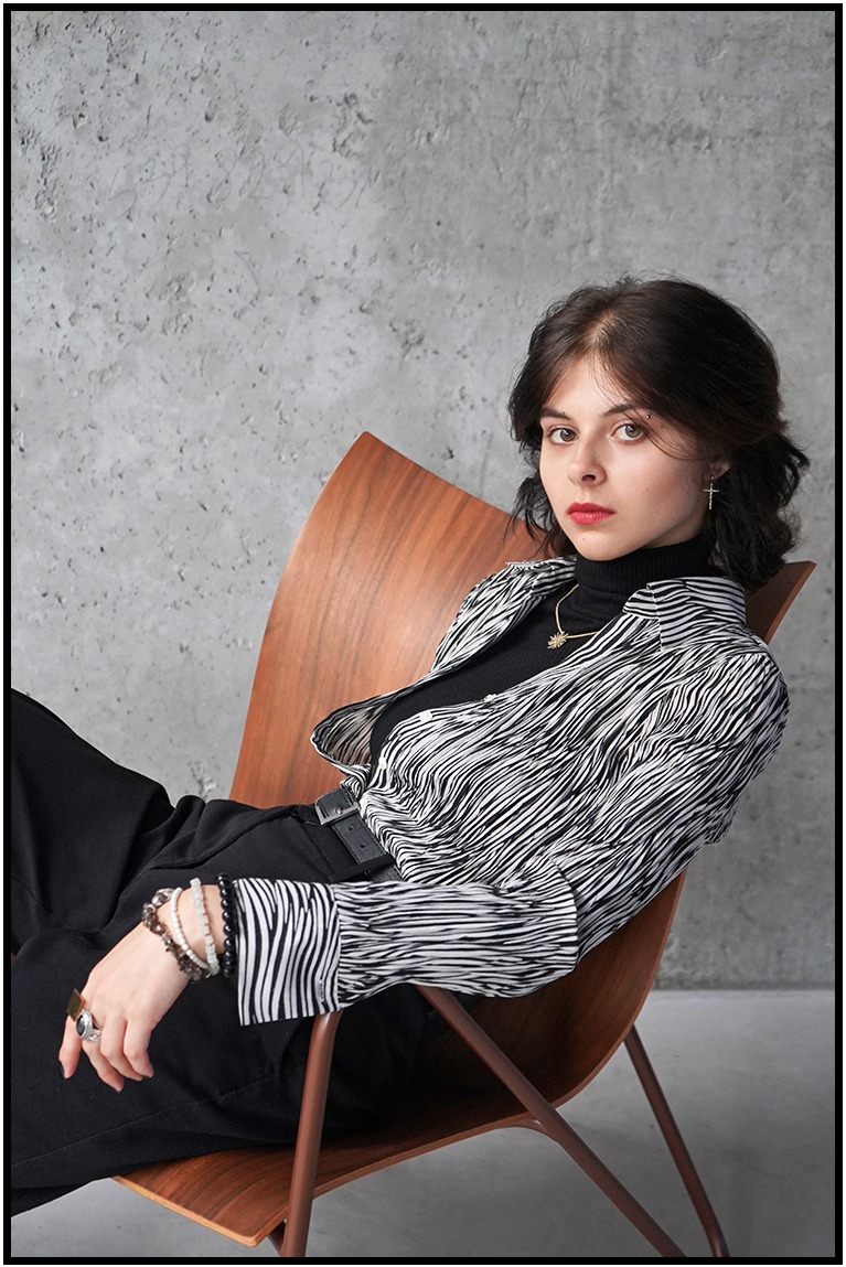 Kobieta w czarno-białej koszuli w paski, siedząca na drewnianym krześle na tle szarej ściany. Uwagę przykuwają detale biżuterii i pierścionki na dłoni.