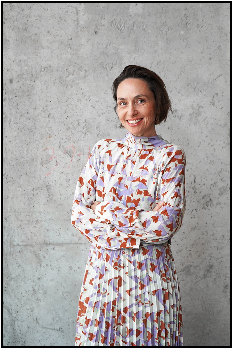 Portret uśmiechniętej kobiety w wzorzystej sukience na tle szarej ściany. Skrzyżowane ramiona, naturalne światło, widoczne detale sukienki i tekstura tła.