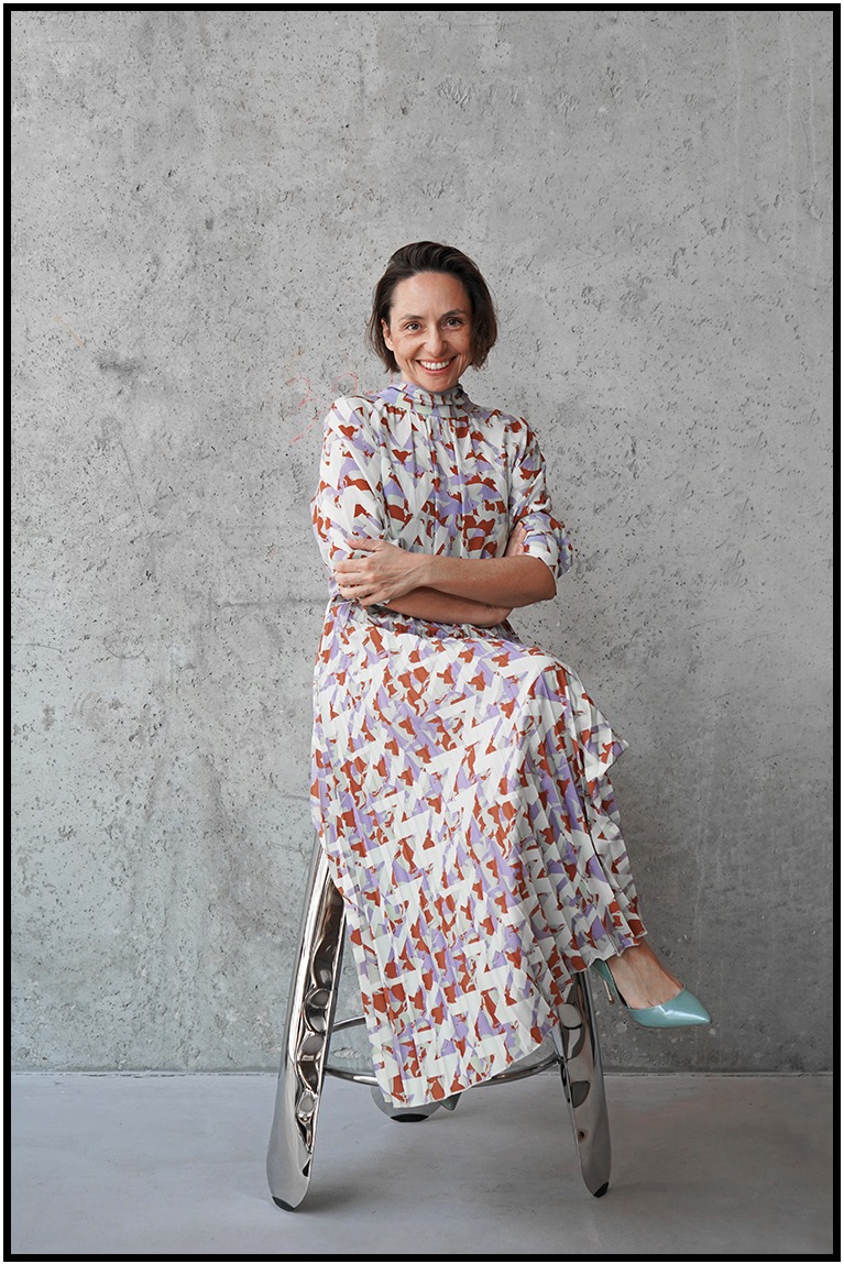 Uśmiechnięta kobieta w wzorzystej sukience siedzi na metalowym stołku na tle surowej, betonowej ściany. Jasne szpilki dodają elegancji.