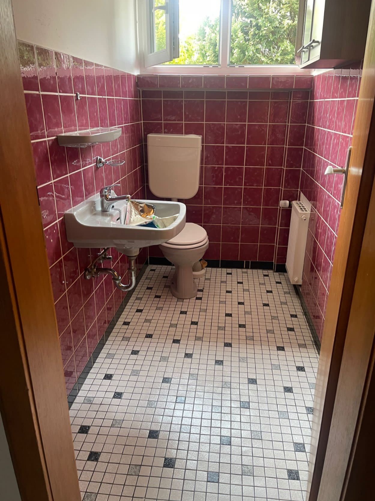 Ukończona łazienka z bordowymi płytkami ściennymi i wzorzystą podłogą. Widoczne umywalka, toaleta i grzejnik. Otwarte okno z widokiem na zieleń.