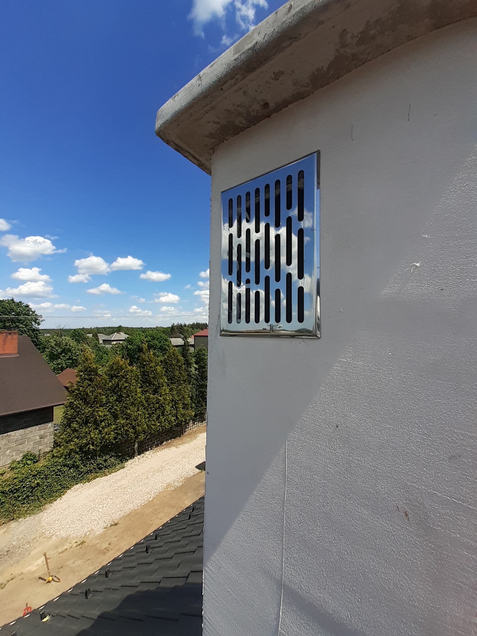 Nierdzewna kratka wentylacyjna zamontowana na otynkowanej ścianie budynku, z widokiem na dach i okoliczną zieleń w tle, w słoneczny dzień.