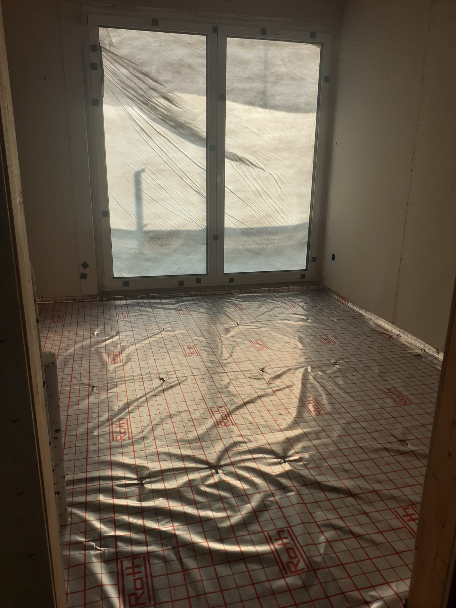 Przygotowanie do ogrzewania podłogowego: folia z siatką na podłożu w pustym pokoju z oknem osłoniętym folią. Widoczne mocowania folii do ściany.