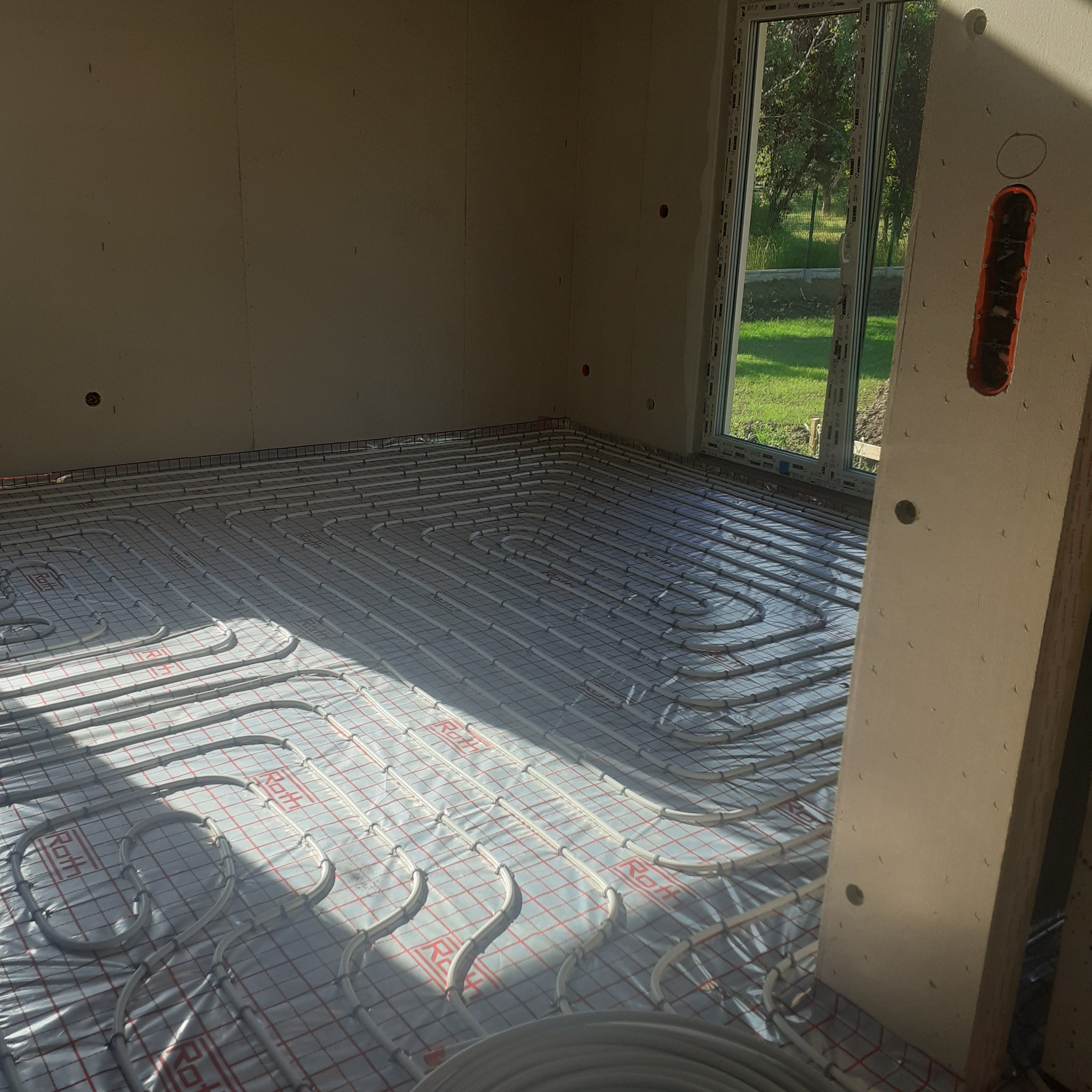 Instalacja ogrzewania podłogowego Roth w jasnym pomieszczeniu z oknem. Widoczne rury na folii z siatką, fragment ściany z gipsu i puszka elektryczna.