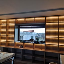 Piotr Skóra - Drewniana zabudowa meblowa z oświetleniem LED, zintegrowana z oknem, widok na budowę za oknem. Na pierwszym planie stół z elementami montażowymi.